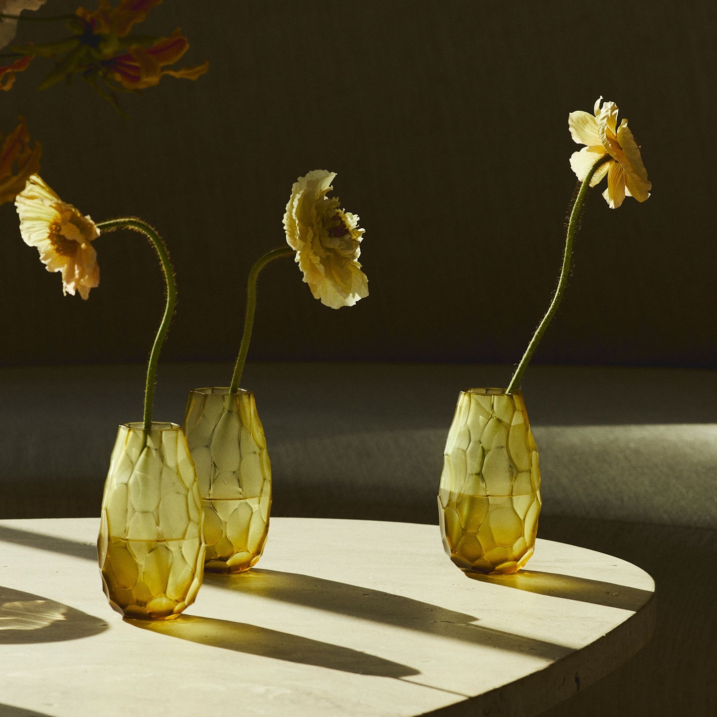 Three textured amber glass vases with a honeycomb pattern, each holding a long-stemmed yellow flower, arranged on a light-colored round table in warm, dramatic sunlight.