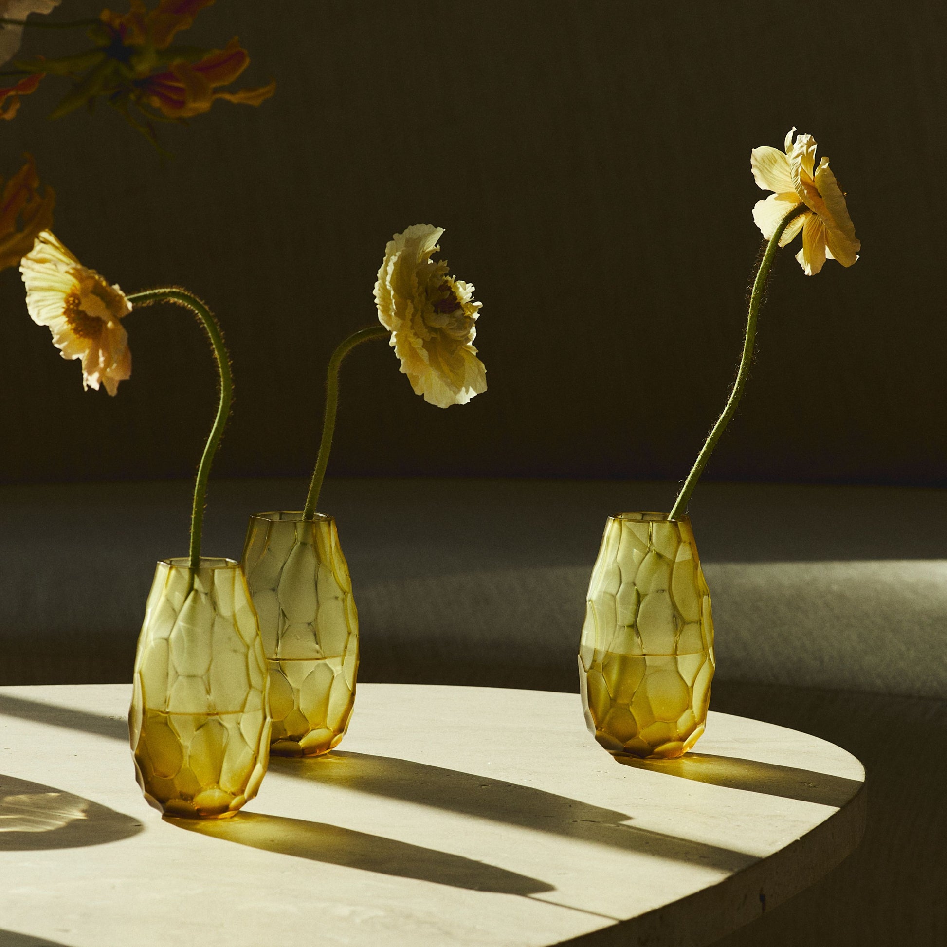 Three textured amber glass vases with a honeycomb pattern, each holding a long-stemmed yellow flower, arranged on a light-colored round table in warm, dramatic sunlight.